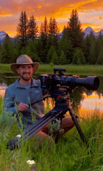 Brandon Navratil Works A Camera In The Field With Mountains And Sunset In The Background